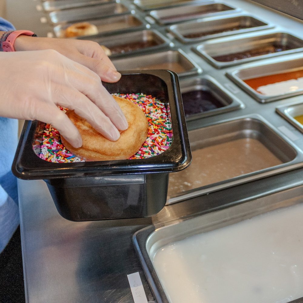 A fresh cake donut being pressed into rainbow sprinkles at the topping station