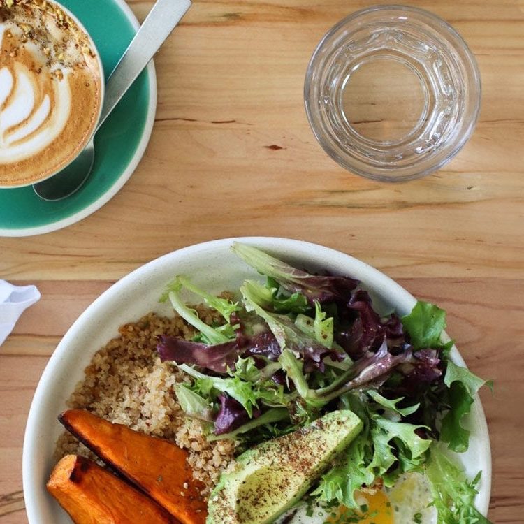 A latte with latte art beside a market bowl of greens, sweet potato and grains