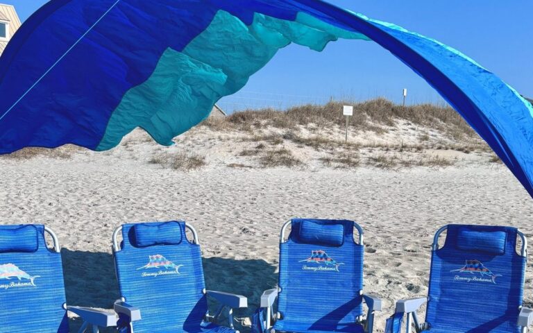 A Shibumi shade over four blue beach chairs in front of the dunes