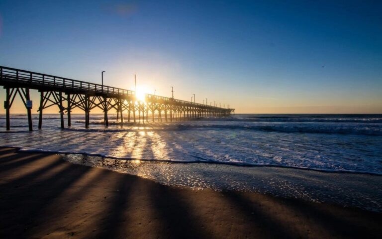 A fishing pier at sunrise with long shadows on the sand