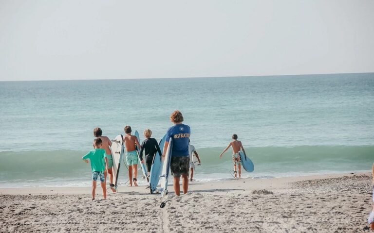 An instructor leading young surfers into the waves, seen from behind